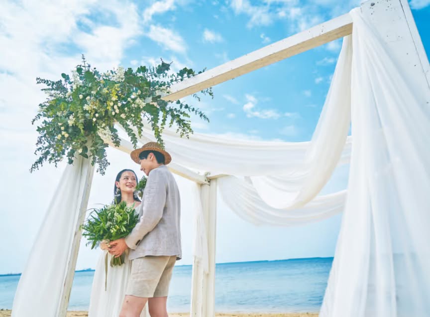 石垣島の澄み渡る青空と海を背景に、白い布とグリーンの装花で飾られたビーチアーチの下で見つめ合う新郎新婦。麦わら帽子を被ったカジュアルな装いの新郎と、ブーケを手に微笑むウエディングドレス姿の花嫁が、リゾートらしい開放感の中で佇む様子。
