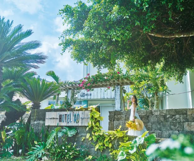 石垣島の「The Beach Terrace HOTEL AO」の木製看板と石垣に囲まれたエントランスにて、黄色のブーケと麦わら帽子を持つウエディングドレス姿の花嫁。南国の植物や鮮やかなブーゲンビリアが咲き誇る、リゾートホテルの入り口での華やかなワンシーン。