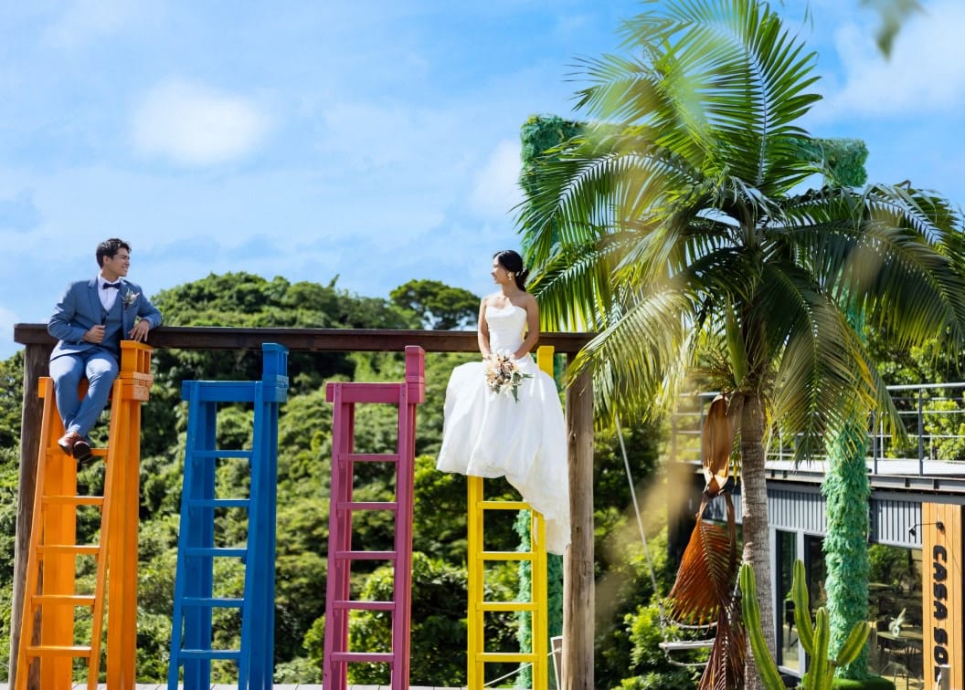 沖縄の青空と豊かな緑の下、カラフルな巨大な椅子の上にそれぞれ座り、楽しそうに見つめ合って微笑む新郎新婦のウエディングフォト