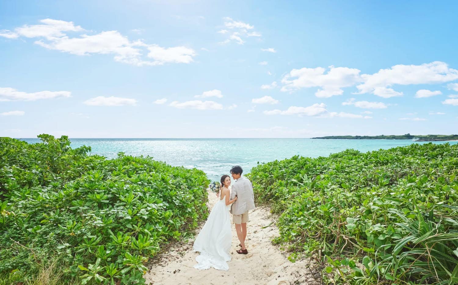 青空とエメラルドグリーンの海を背景に、白い砂浜の小道を歩くウエディングドレス姿の新婦とカジュアルなスーツの新郎。緑豊かな植物に囲まれたロケーションで、二人が寄り添いながら笑顔で振り返るビーチフォトウエディングの様子。