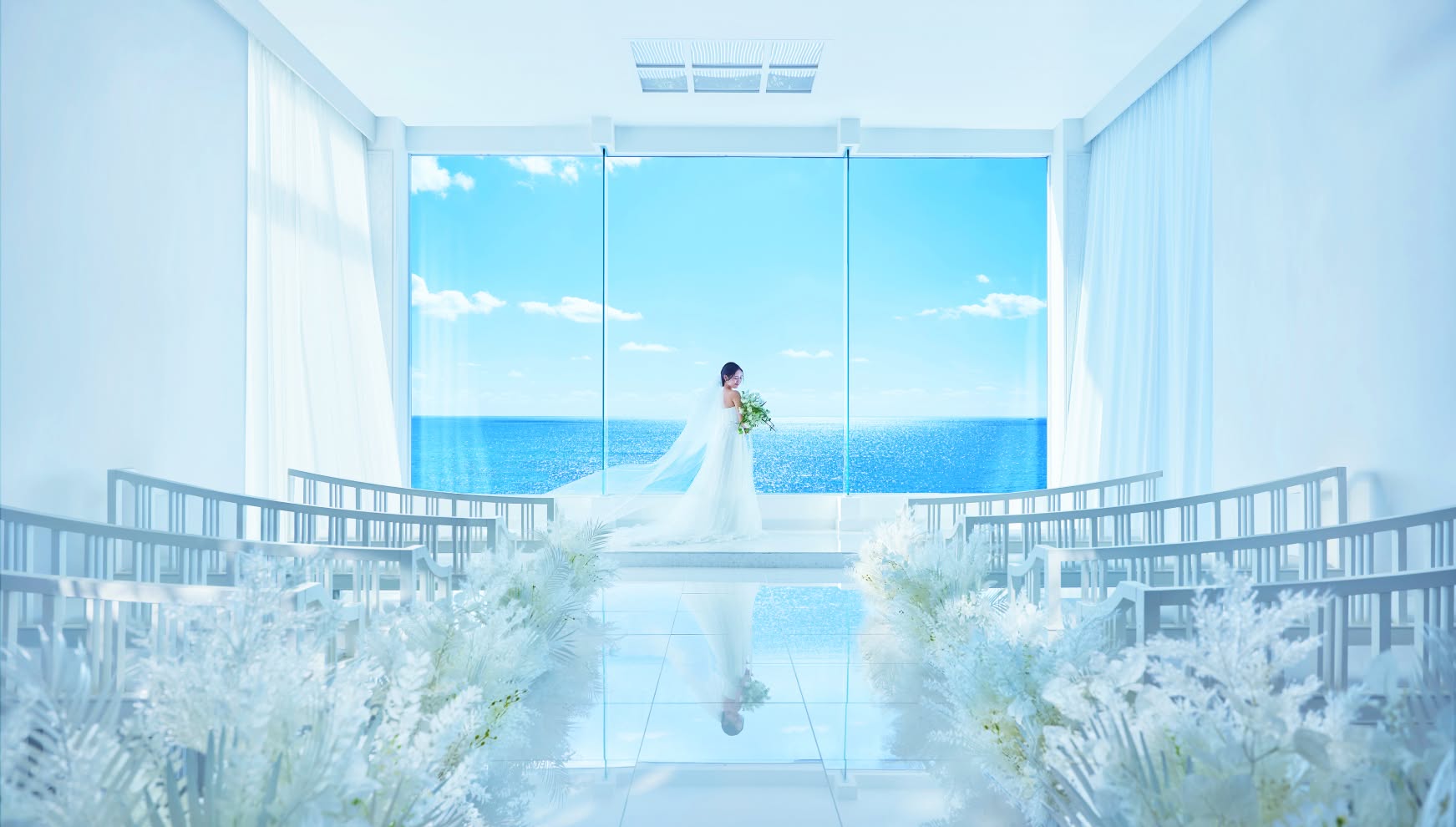 青い海が一望できる全面ガラス張りの白亜のチャペルに、一人佇むウエディングドレス姿の花嫁。高い透明感と自然光が美しい空間で、バージンロードの装花がリゾートウエディングの清らかな雰囲気を引き立てています。