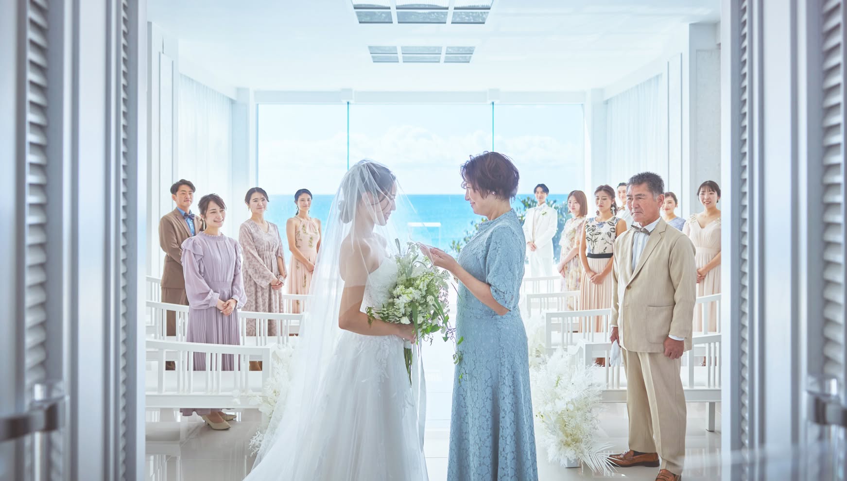 海が見える開放的なチャペルで、ゲストに見守られながら母親からベールダウンを受ける花嫁。家族の絆を感じる感動的な挙式シーンで、白を基調とした会場にリゾートらしい青い海と空が美しく映えています。