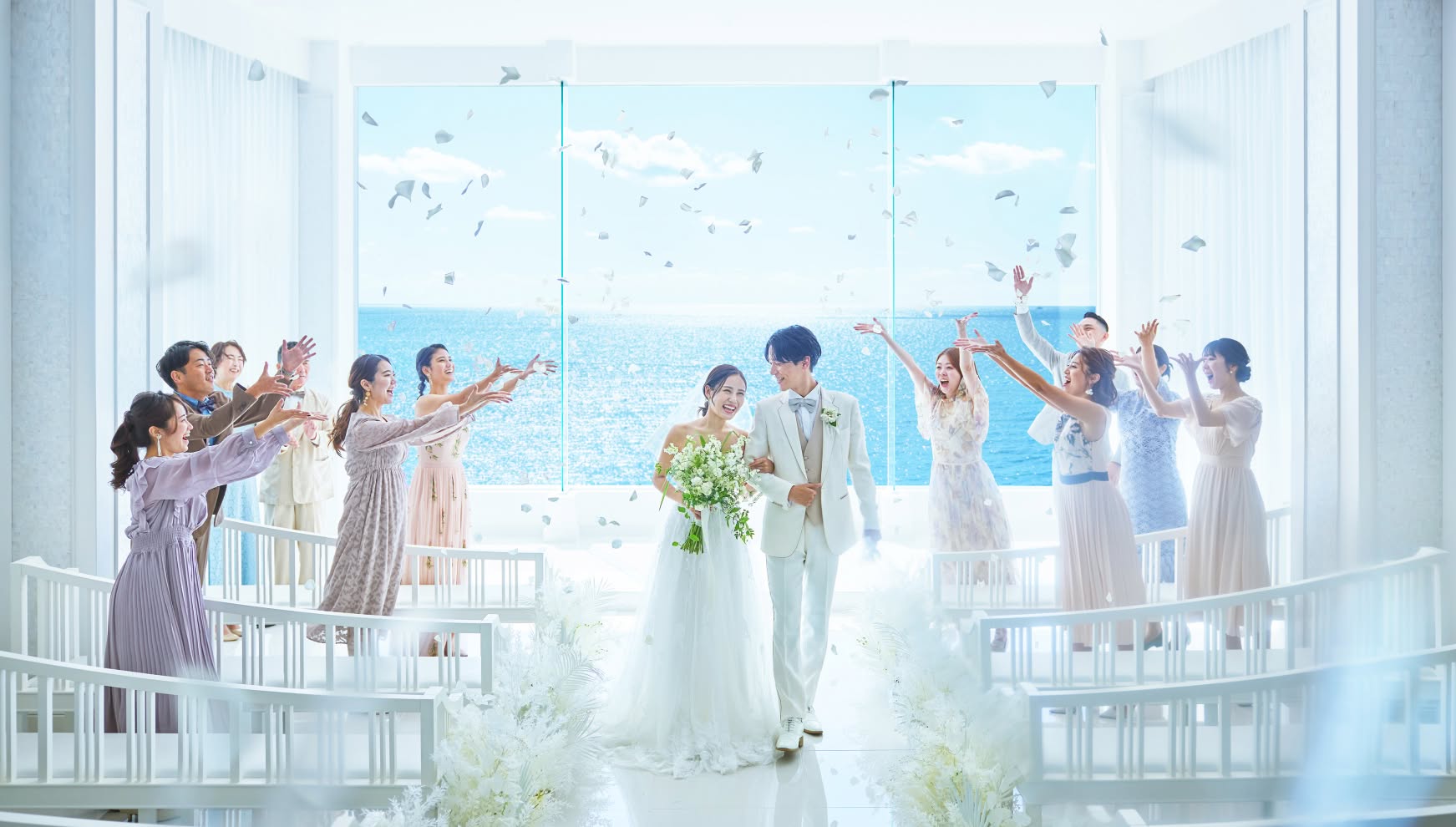 海が見える白亜のチャペルで、ゲストからフラワーシャワーの祝福を受ける笑顔の新郎新婦。青い空と海が広がる開放的な空間で、バージンロードを歩む二人の幸せ溢れるリゾートウエディングの退場シーンです。
