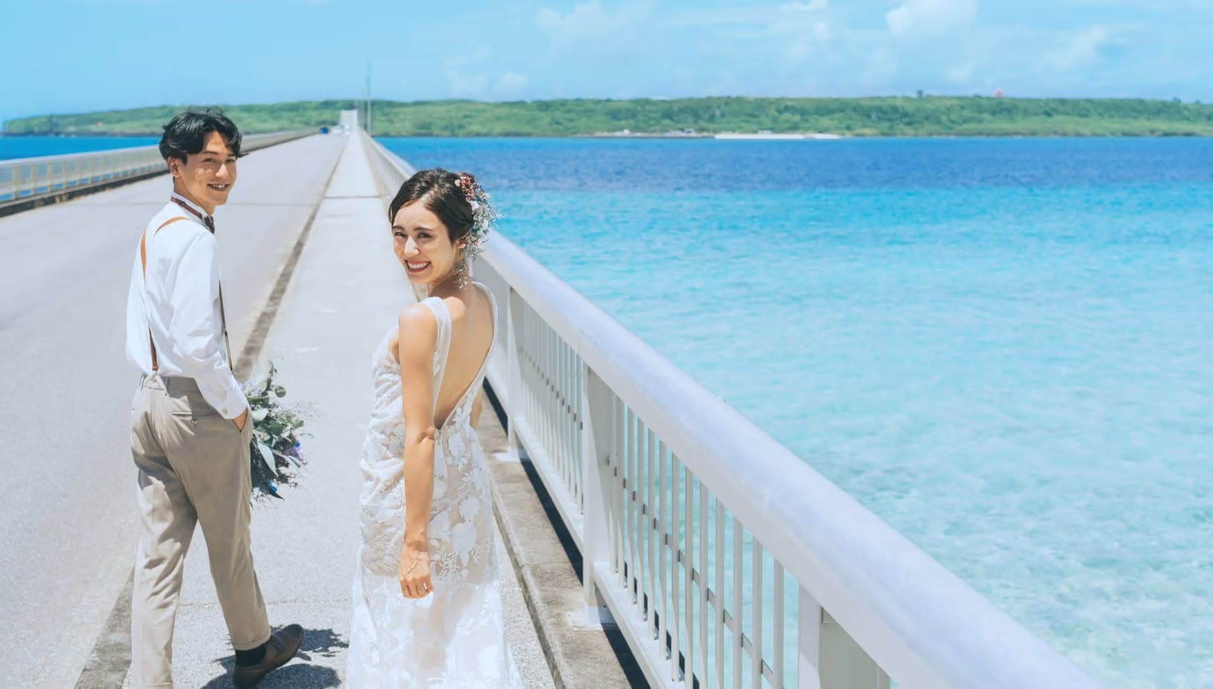 エメラルドグリーンの海へと続く橋の上で、笑顔で振り返る新郎新婦。沖縄の美しい絶景をバックにしたフォトツアーの一コマで、リゾートウエディングならではの開放感溢れるロケーションフォトシーンです。