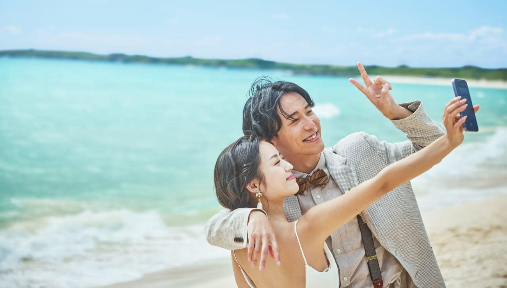 美しいエメラルドグリーンの海をバックに、ビーチで自撮りを楽しむ笑顔の新郎新婦。自然体な二人の表情が印象的な、沖縄のリゾートウエディングにおけるフォトツアーでの楽しげなオフショットです。