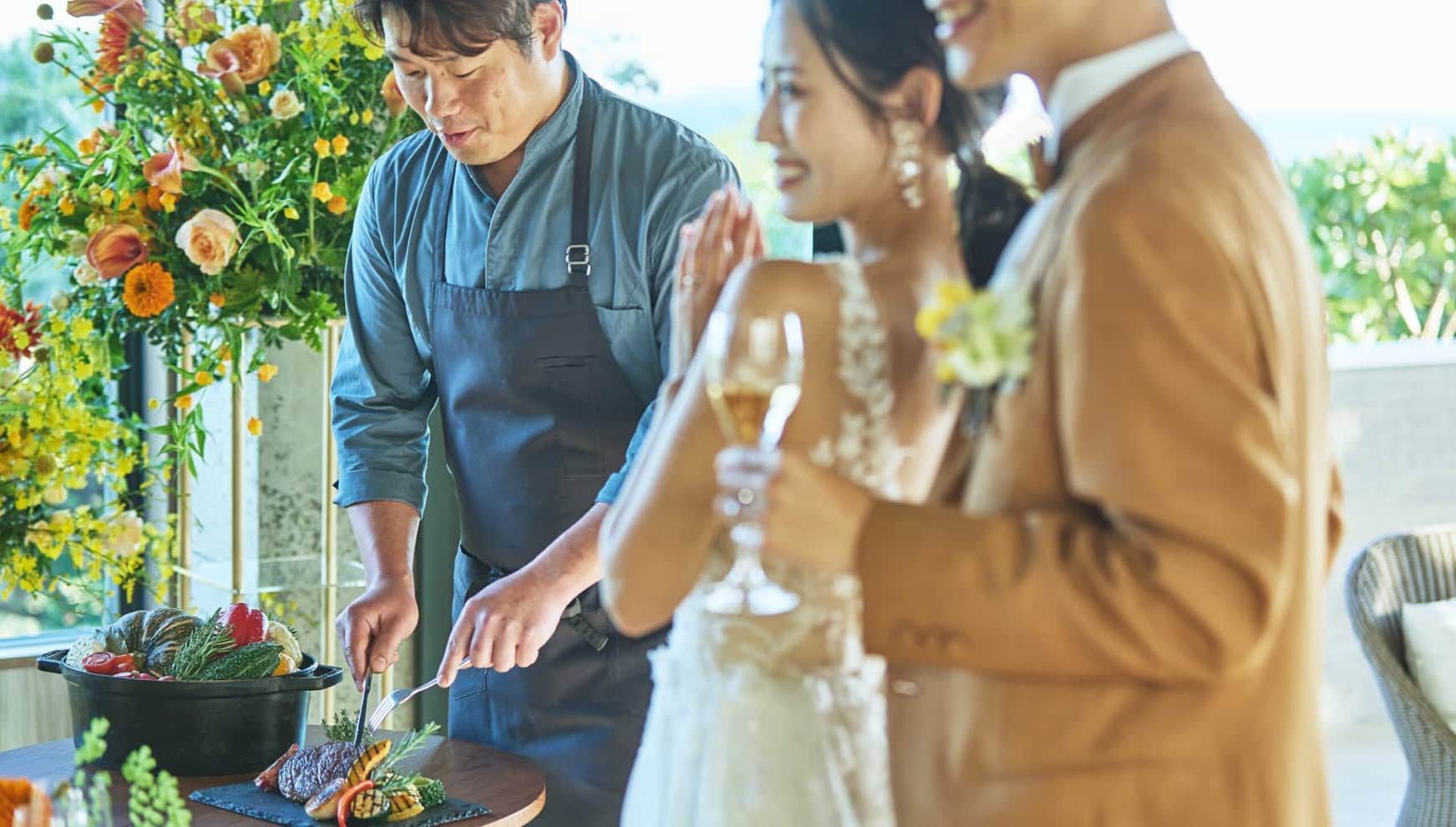 リゾートウエディングのパーティー会場で、新郎新婦の目の前で肉料理を切り分けるシェフ。ゲストへのおもてなしを感じるライブキッチン演出の一コマで、色鮮やかな夏野菜や華やかな装花がパーティーを彩っています。