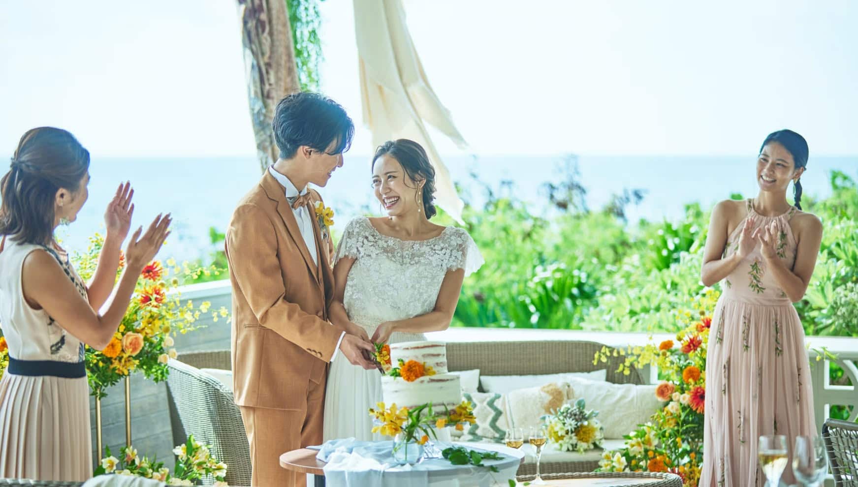 海を一望する開放的なテラスで、ゲストの拍手に包まれながらウエディングケーキ入刀を行う新郎新婦。オレンジの花々で彩られたナチュラルなケーキと、リゾートの心地よい風を感じる和やかなウエディングパーティーのワンシーンです。