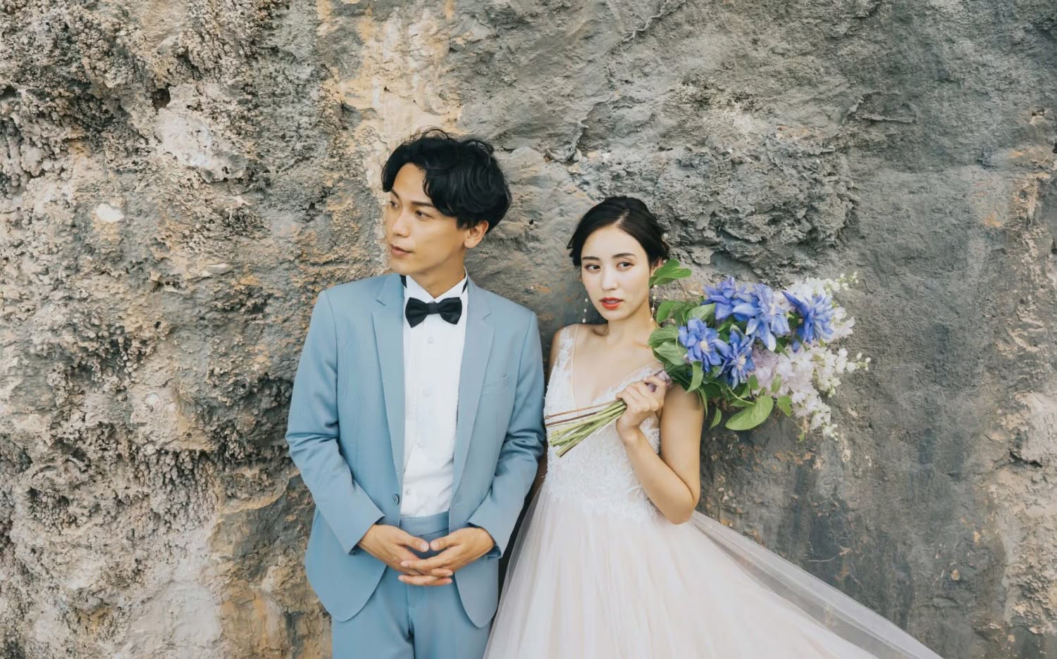 ゴツゴツとした岩肌を背景に、ブルーのタキシードを着た新郎と、青いブーケを手にしたウエディングドレス姿の新婦が並ぶカット。洗練された雰囲気の中で自然の質感を活かした、ドラマチックなロケーションフォトウエディング。