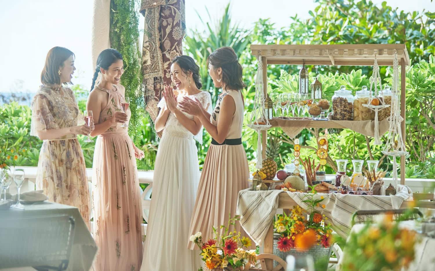 緑豊かな屋外会場で、ドリンクを手に談笑するウエディングドレス姿の新婦と3人のブライズメイド。ナチュラルな装飾のフードカートや色鮮やかな花々に囲まれた、アットホームで華やかな結婚式二次会パーティーの様子。