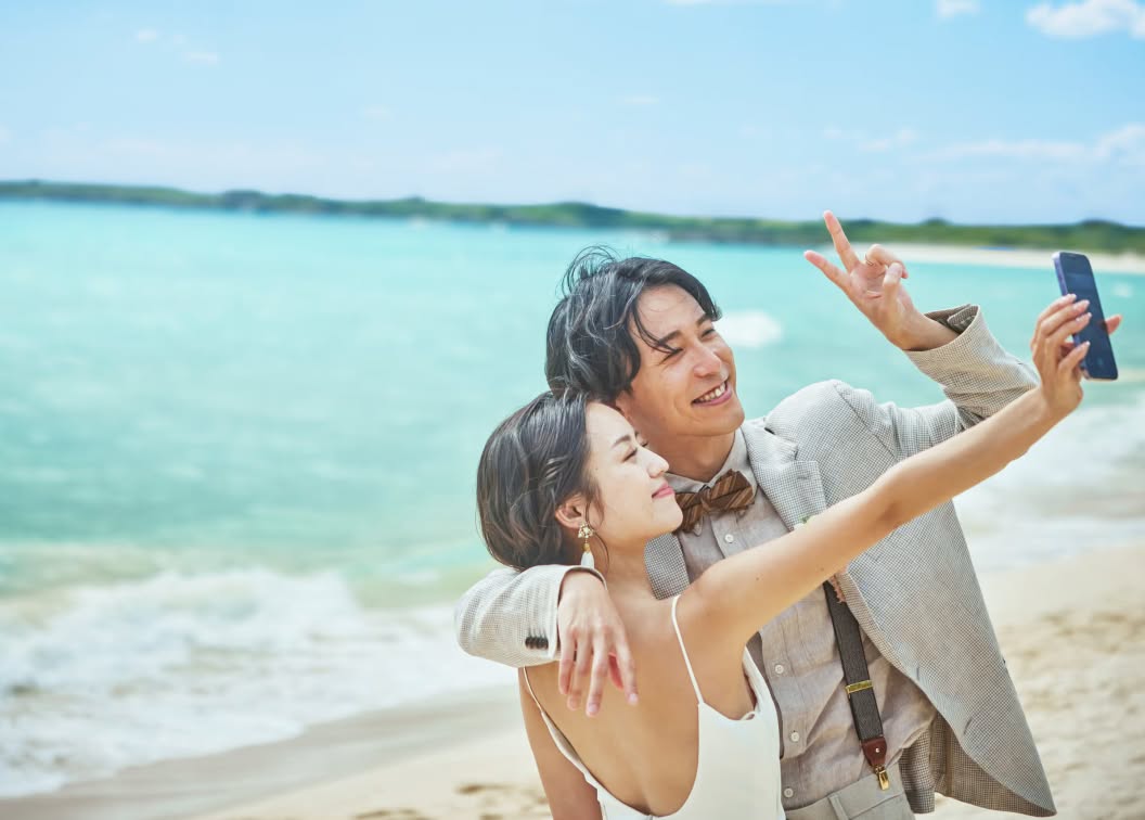 美しいエメラルドグリーンの海をバックに、ビーチで自撮りを楽しむ笑顔の新郎新婦。自然体な二人の表情が印象的な、沖縄のリゾートウエディングにおけるフォトツアーでの楽しげなオフショットです。