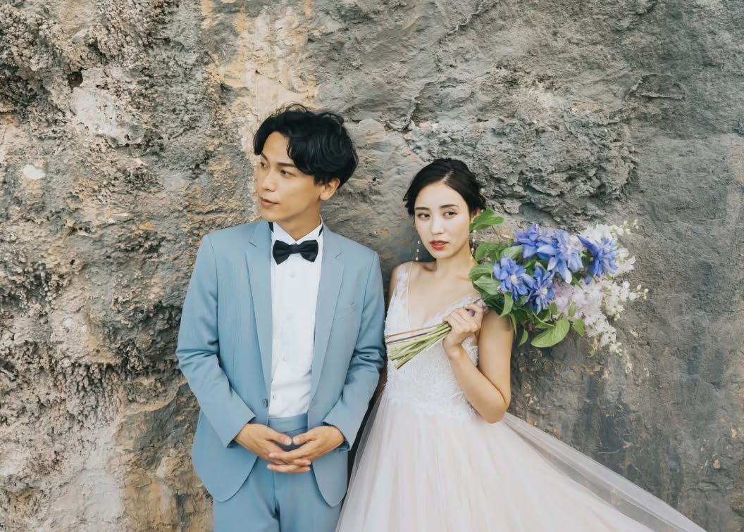 ゴツゴツとした岩肌を背景に、ブルーのタキシードを着た新郎と、青いブーケを手にしたウエディングドレス姿の新婦が並ぶカット。洗練された雰囲気の中で自然の質感を活かした、ドラマチックなロケーションフォトウエディング。