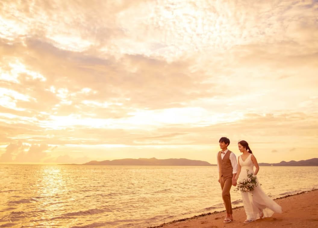 夕暮れ時のビーチを歩く新郎新婦のロケーションフォト。オレンジ色に染まる海と空を背景に、ウエディングドレス姿の花嫁とベージュのスーツを着た新郎が手をつなぐ、開放感あふれるビーチウエディングのワンシーンです。
