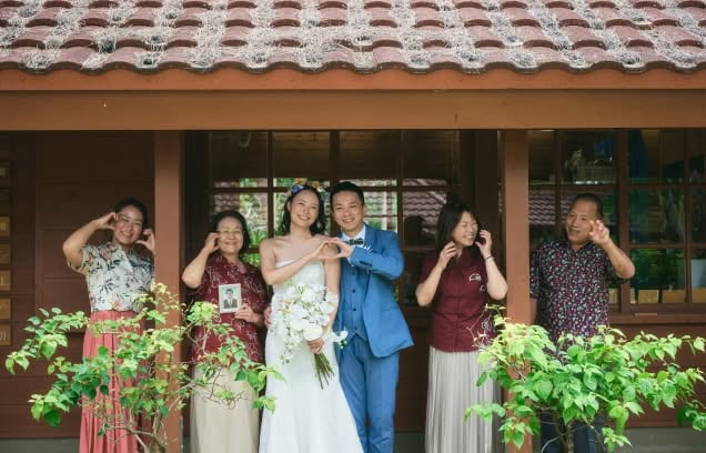 沖縄の木造建築の前で、白いドレスのウエディング姿の新婦と青いスーツの新郎を家族が囲む記念写真。全員でハートやポーズを作り、笑顔で並んでいます。赤瓦の屋根と緑の植栽が南国らしい温かな雰囲気を演出する、アットホームな結婚式の様子です。