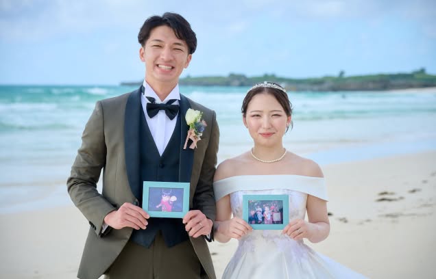 沖縄のビーチで、子供時代の写真が入ったフォトフレームを手にするウエディング姿の新郎新婦。エメラルドグリーンの海を背景に、タキシードとオフショルダーのドレスを纏った二人が笑顔で並ぶ、リゾート婚のメモリアルなワンシーンです。