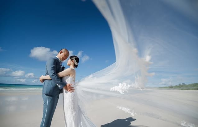 沖縄の白い砂浜で、長いロングベールを風になびかせて見つめ合うウエディング姿の新郎新婦。青い空と海をバックに、総レースのドレスとブルーのスーツが映える、ドラマチックで開放感あふれるビーチフォトのワンシーンです。