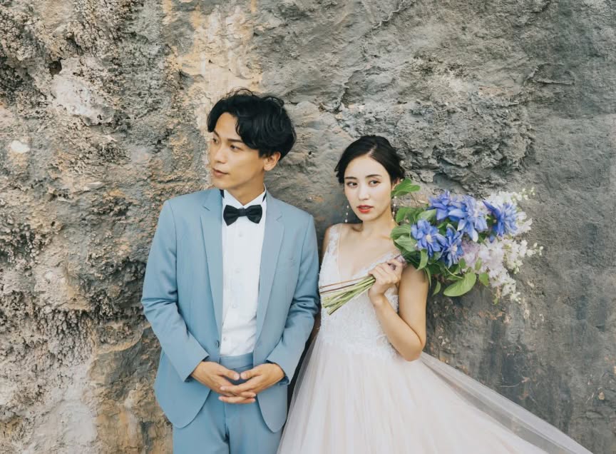 ゴツゴツとした岩肌を背景に、ブルーのタキシードを着た新郎と、青いブーケを手にしたウエディングドレス姿の新婦が並ぶカット。洗練された雰囲気の中で自然の質感を活かした、ドラマチックなロケーションフォトウエディング。