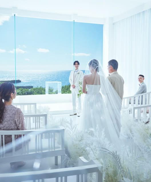 青い海が一望できる真っ白なチャペルで、新婦の入場を待つ新郎の姿。ゲストに見守られながら、バージンロードを歩むウエディングドレス姿の花嫁と父親。リゾートウエディングの挙式シーンです。