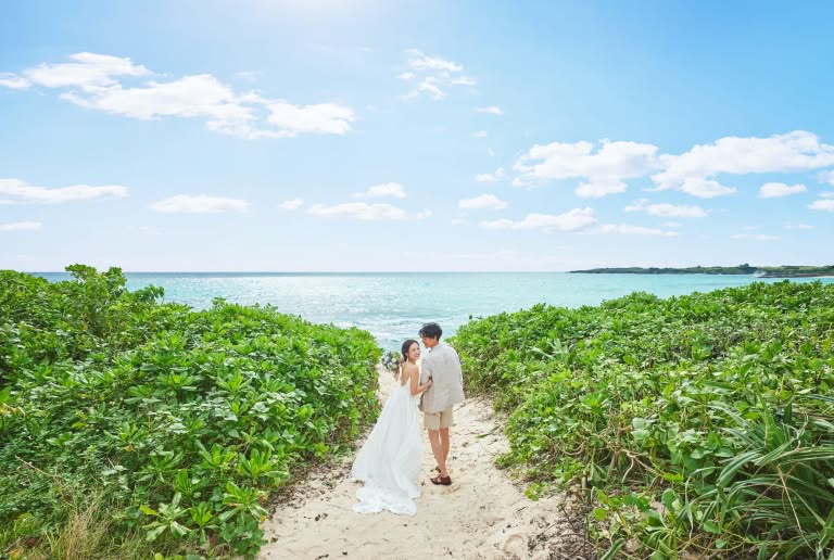 青空とエメラルドグリーンの海を背景に、白い砂浜の小道を歩くウエディングドレス姿の新婦とカジュアルなスーツの新郎。緑豊かな植物に囲まれたロケーションで、二人が寄り添いながら笑顔で振り返るビーチフォトウエディングの様子。