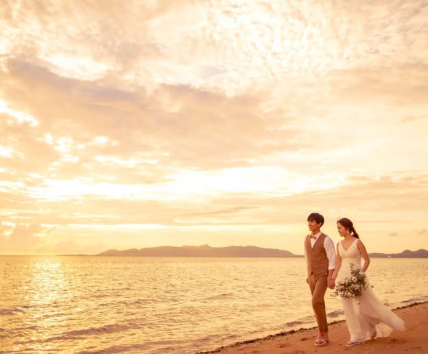 夕暮れ時のビーチを歩く新郎新婦のロケーションフォト。オレンジ色に染まる海と空を背景に、ウエディングドレス姿の花嫁とベージュのスーツを着た新郎が手をつなぐ、開放感あふれるビーチウエディングのワンシーンです。