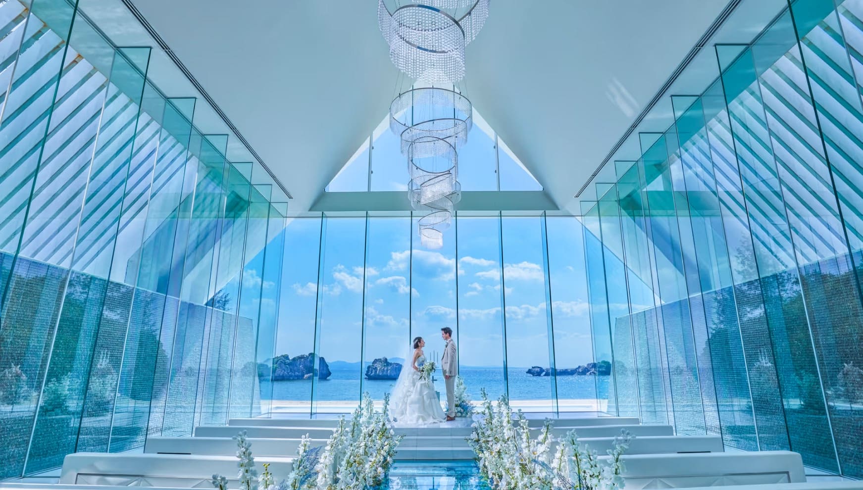 「アイネス ヴィラノッツェ 沖縄」の全面ガラス張りのチャペルにて、祭壇に立つ新郎新婦。天井から吊るされた螺旋状のクリスタルシャンデリアと、背景に広がる沖縄の青い海が一体となった、幻想的で透明感あふれる挙式会場の風景。