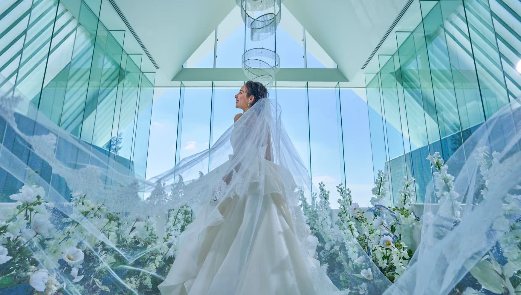 「アイネス ヴィラノッツェ 沖縄」のガラス張りのチャペルにて、長いベールを美しくなびかせるウエディングドレス姿の新婦のバックスタイル。天井から降り注ぐクリスタルと白い花々に囲まれ、青空を見上げる幻想的でドラマチックなワンシーン。
