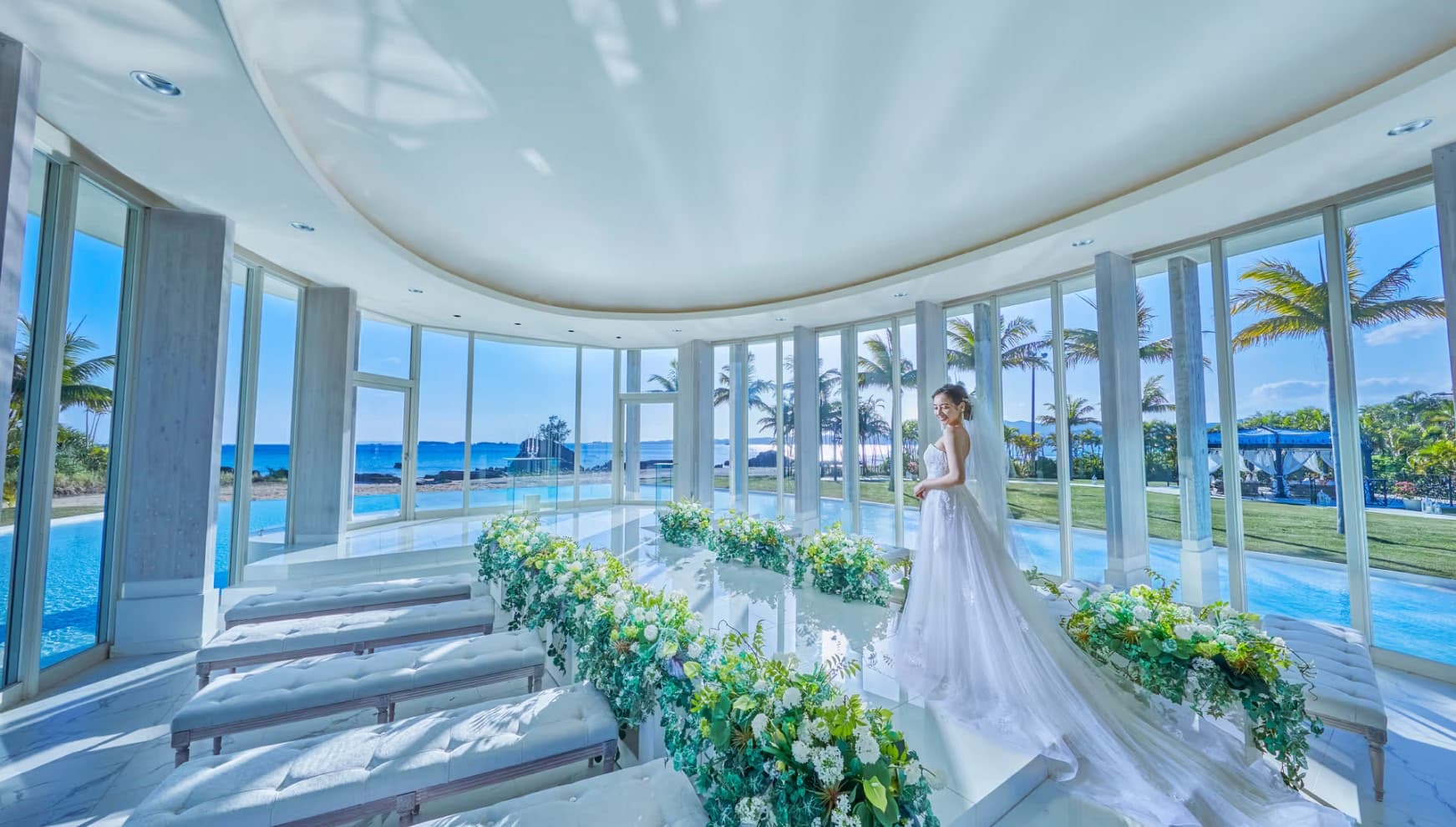 海が見える全面ガラス張りの開放的なチャペルで、ウエディングドレス姿の女性が海を眺めて佇む様子。大理石のバージンロードの両脇には緑と白の花々が飾られ、南国のヤシの木と青い空が広がるリゾート感溢れる空間。
