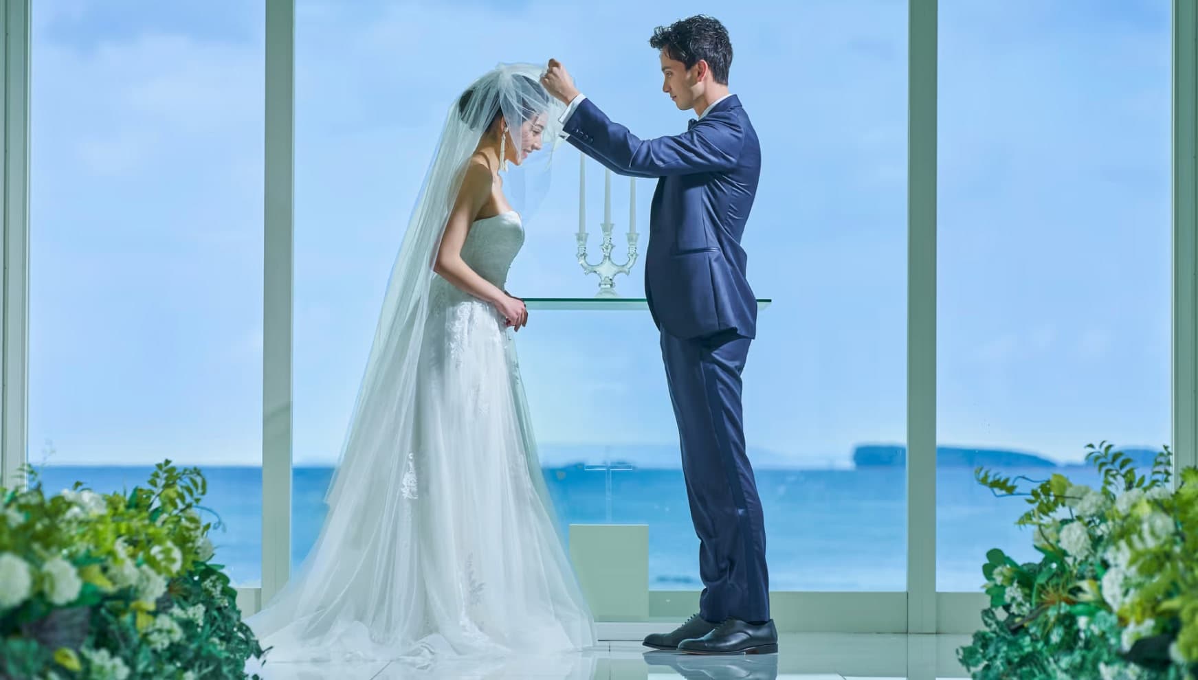 海の見える開放的なチャペルで、タキシード姿の新郎がウエディングドレス姿の新婦のベールアップを行う儀式のシーン。背景には青い海と空が広がり、手前には装飾の花々が添えられた、リゾート感溢れる幻想的な雰囲気。