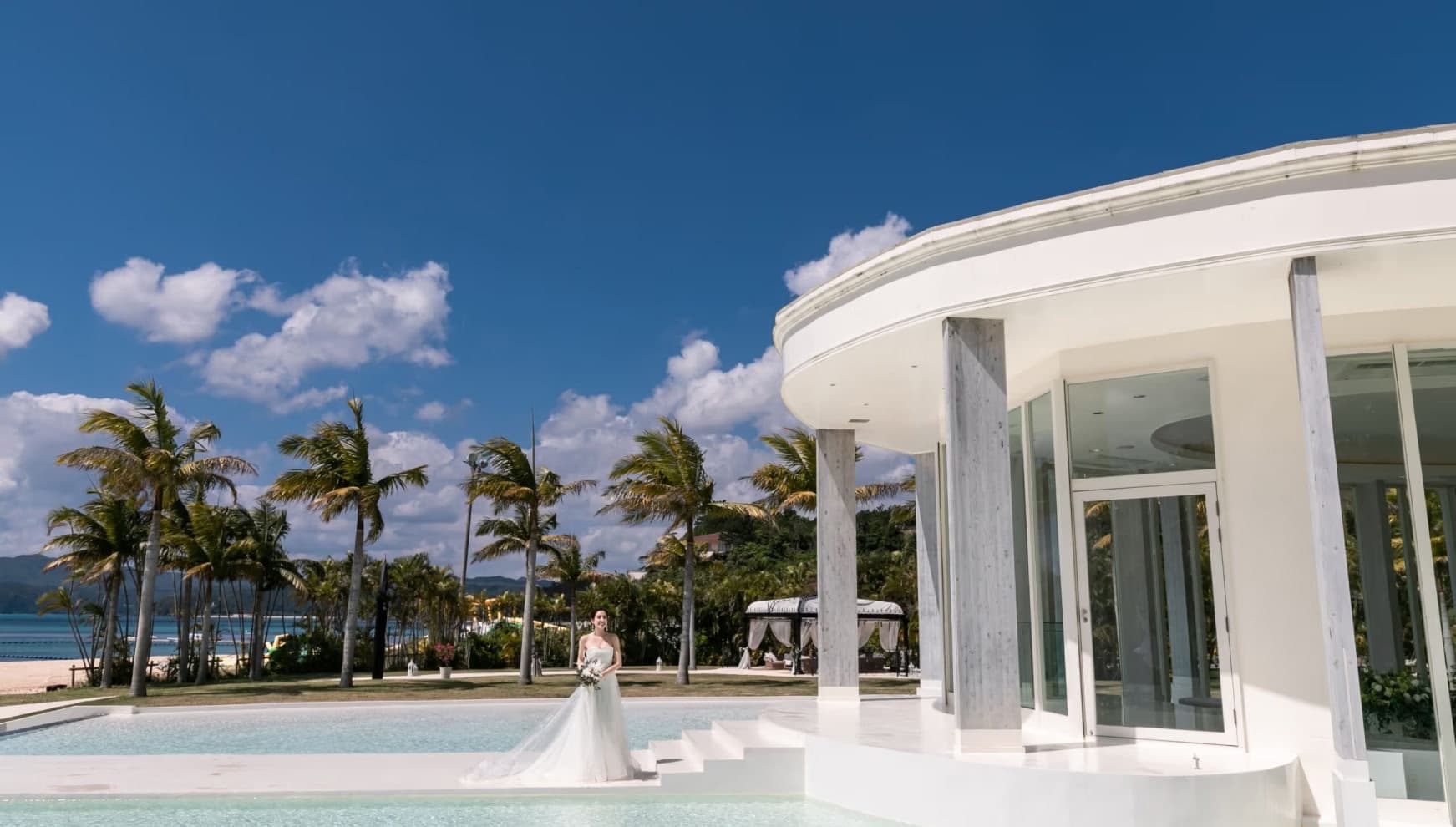 青空の下、白い円形のモダンなチャペル外観とプールサイドに佇むウエディングドレス姿の新婦。背景には多くのヤシの木とエメラルドグリーンの海が広がり、南国リゾートの開放感あふれるロケーションフォトの様子。