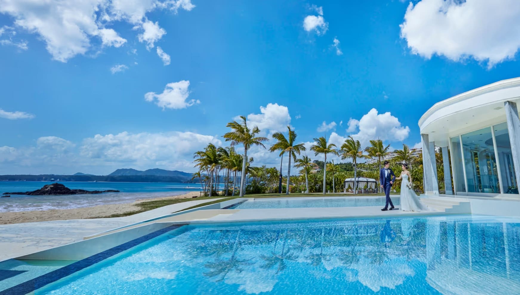 青い空と海を背景に、プールサイドを仲睦まじく歩くウエディングドレス姿の新婦とタキシード姿の新郎。ヤシの木が並ぶリゾート地で、水面に景色が反射する美しいロケーションの中でのウエディングフォトシーン。