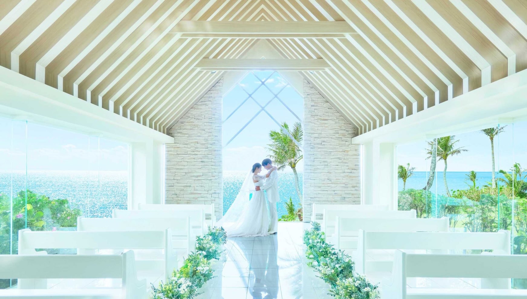 「ムーンビーチウェディング」のチャペルにて、祭壇の前で見つめ合う白いタキシードの新郎とウエディングドレスの新婦。木目調の温かみがある高い天井と、正面のガラス越しに広がる青い海と空、ヤシの木がリゾート感を演出する挙式会場。