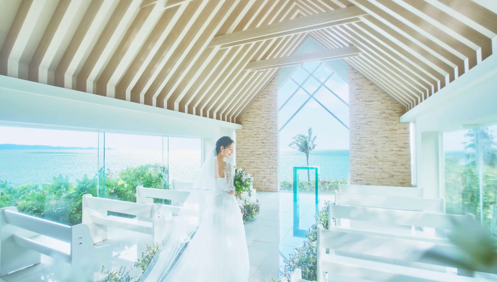 光溢れるチャペルにて、ブーケを手に祭壇前で佇むウエディングドレス姿の新婦。木組みの天井と石造りの壁、大きな窓から一望できる青い海が調和した、開放感あふれる挙式会場でのバックショット。