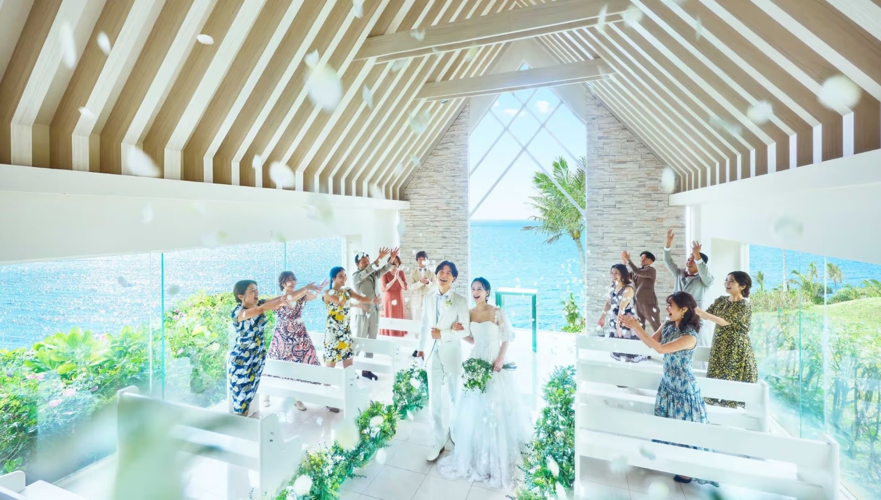 開放的なチャペルにて、ゲストからのフラワーシャワーに包まれる白いタキシードの新郎とウエディングドレスの新婦。木組みの天井と大パノラマの海が広がる空間で、参列者の笑顔に祝福される幸せなウエディングシーン。