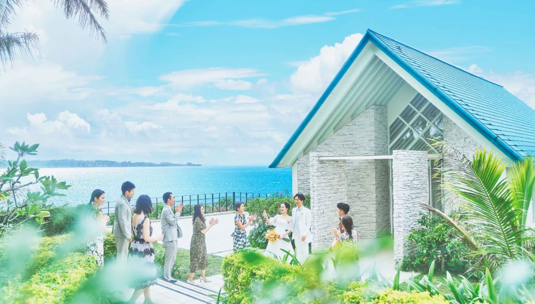 青い屋根が特徴的なチャペル外観。海を一望する絶景のロケーションで、ウエディングドレス姿の新婦と白いタキシードの新郎が、ゲストに囲まれ祝福を受けるリゾートウエディングのワンシーン。