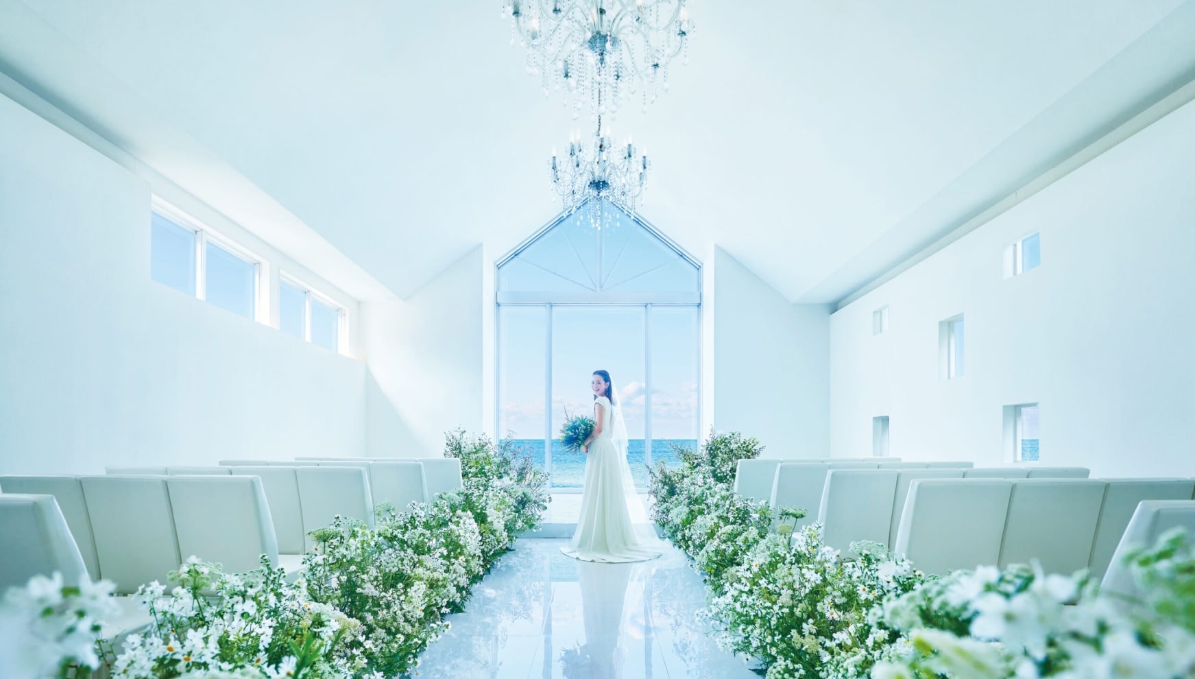 ザ・ビーチテラス ホテルアオ石垣の白亜のチャペル内観。シャンデリアが輝く高い天井と、正面の大きな窓から見える石垣島の青い海。バージンロードに飾られた白い花々に囲まれ、ウエディングドレス姿の新婦が微笑む幻想的なシーン。