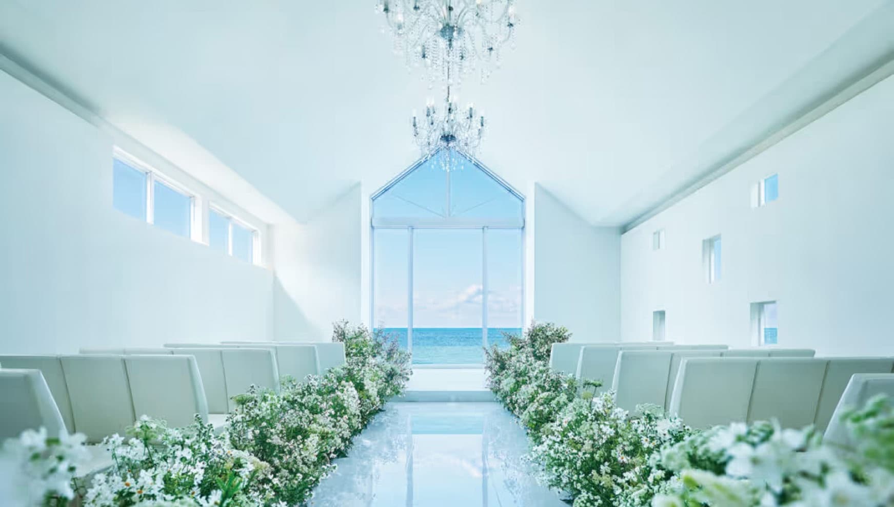 ザ・ビーチテラス ホテルアオ石垣の白亜のチャペル内観。シャンデリアが輝く高い天井と、正面の大きな窓から一望できる石垣島の青い海。バージンロード沿いに白い花々が豊かに飾られた、開放感あふれる挙式会場の全景。