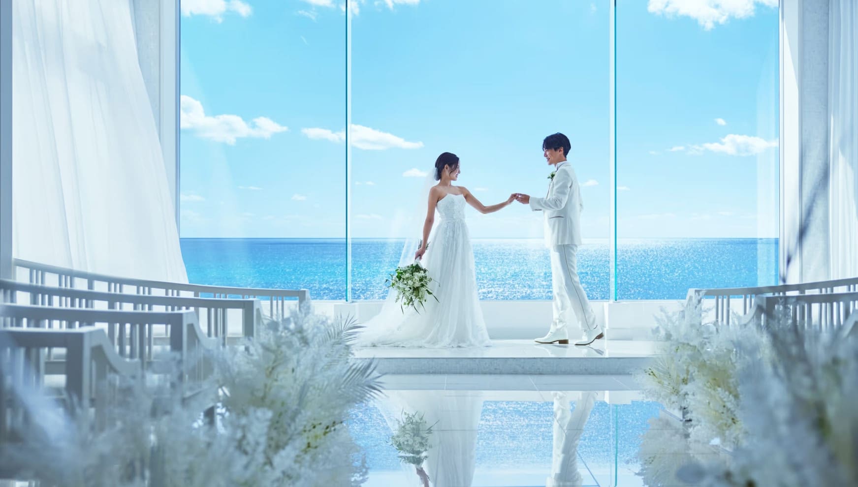 ザ・サーフ シーサイドシーン宮古島の光溢れるチャペルで、手を取り合い見つめ合うウエディングドレス姿の新婦と白いタキシード姿の新郎。背景の大きな窓には宮古島の青い海が広がり、床面に景色が反射する幻想的なワンシーン。
