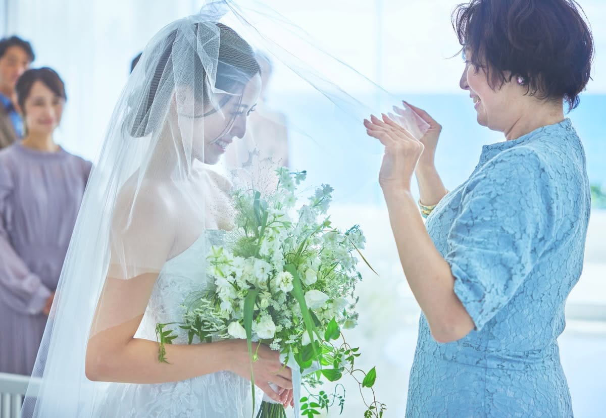 チャペルにて、母親からベールダウンを受ける新婦のアップ。透き通るようなベールの向こうで微笑むウエディングドレス姿の新婦と、慈しみ深く見守る母親の姿が感動的な挙式前のワンシーン。