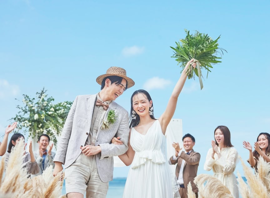 青い海を背景にしたビーチウエディングで、幸せそうな新郎新婦と、二人を笑顔で見守る参列者たち
